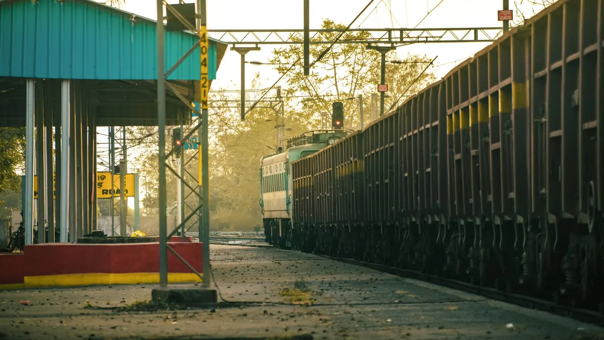 A freight train arrives at an Indian railway station platform during a calm day.