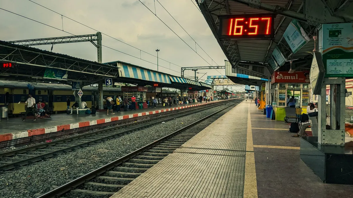 An urban railway station platform at 16:57 with digital clock and passengers in waiting.