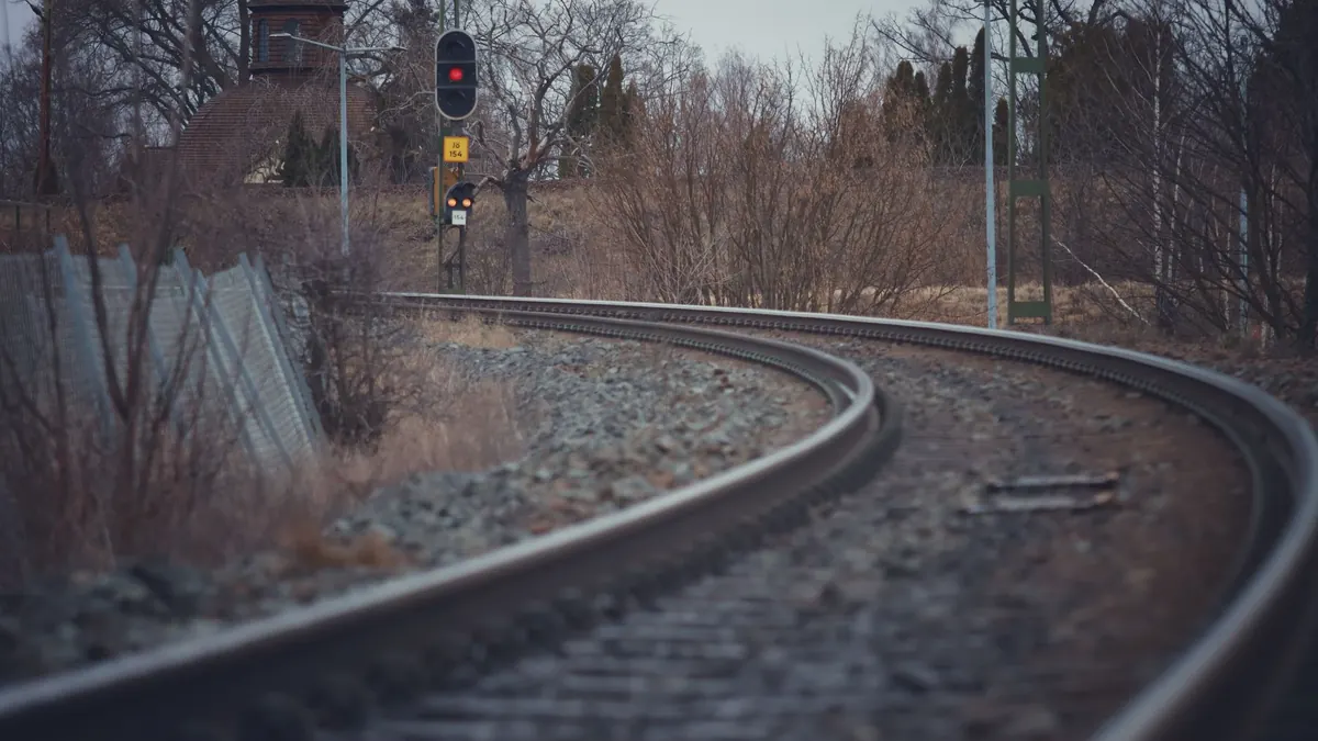 A scenic view of a railway track curving through a landscape with bare trees and a distant traffic light in winter.