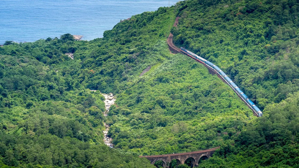 From above of train driving on railroad between lush trees on ridge against sea in daytime